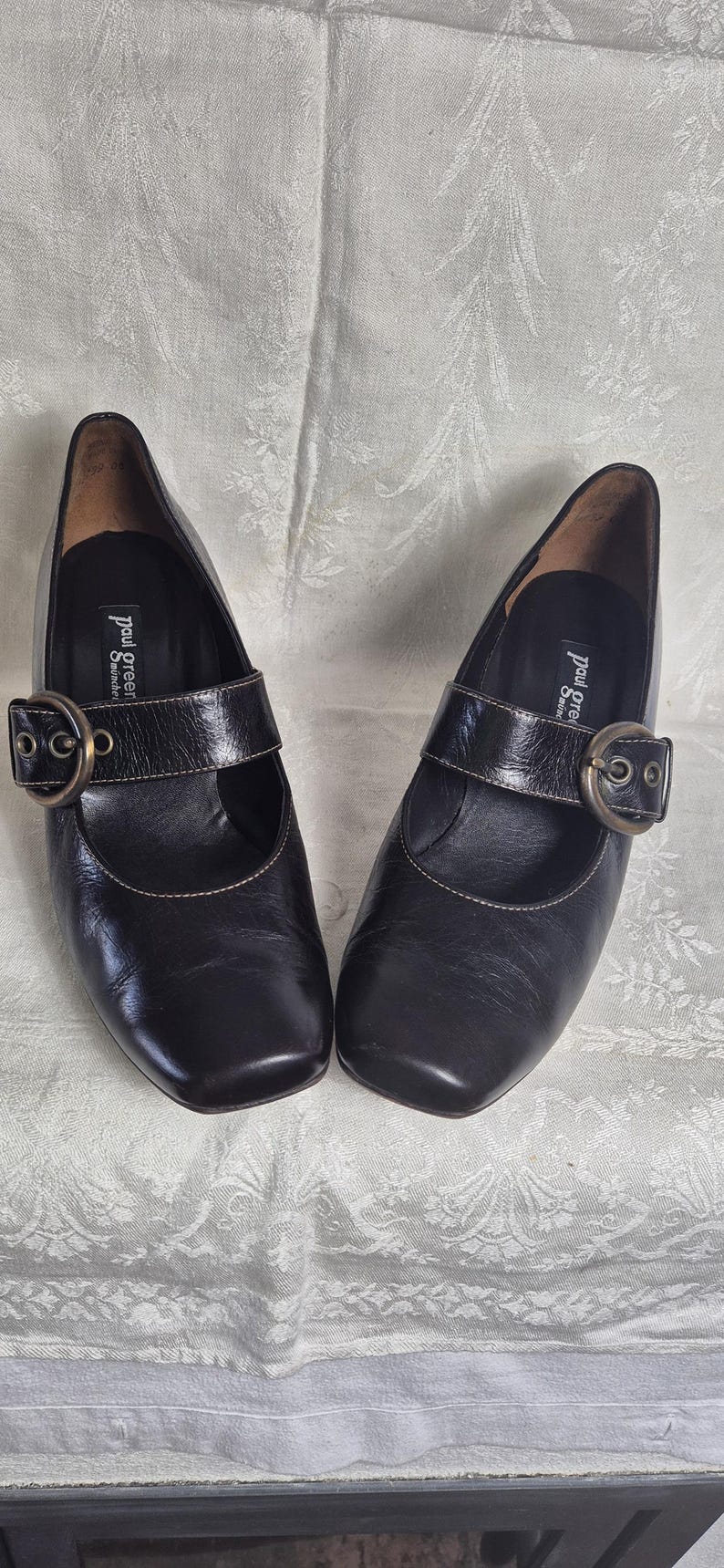 May include: A pair of black leather Mary Jane style shoes with a square toe and a buckle closure. The shoes have a small heel and are displayed on a patterned surface. The brand name "paul green" is visible on the insole.