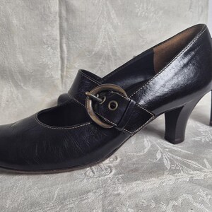 May include: A pair of black leather Mary Jane style shoes with a buckle closure and a small heel. The shoes have a rounded toe and a strap that fastens across the top of the foot. The heel height is approximately 2.5 inches.