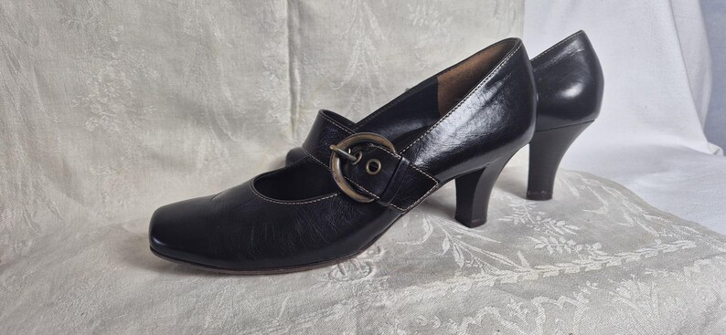 May include: A pair of black leather Mary Jane style shoes with a buckle closure and a small heel. The shoes have a rounded toe and a strap that fastens across the top of the foot. The heel height is approximately 2.5 inches.