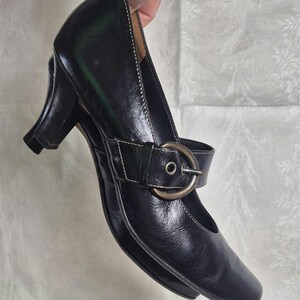 May include: Black leather Mary Jane style shoe with a small heel. The shoe features a strap across the top with a buckle closure. White stitching accents the edges of the shoe.