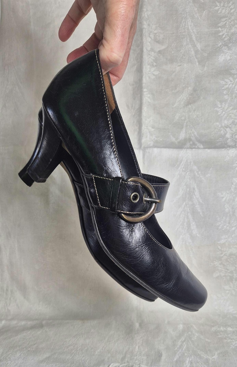 May include: Black leather Mary Jane style shoe with a small heel. The shoe features a strap across the top with a buckle closure. White stitching accents the edges of the shoe.