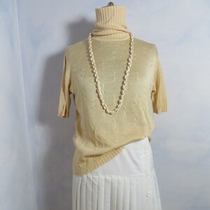Op de afbeelding: Een beige gebreide trui met een rolkraag en korte mouwen. De trui wordt gedragen over een witte geplooide rok. Een witte schelpenketting wordt om de nek gedragen.