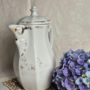 May include: White porcelain pitcher with a lid, featuring gold accents and floral patterns. The pitcher sits on a small, embroidered coaster. A bouquet of purple hydrangeas is next to the pitcher. A vintage-style decorative item.