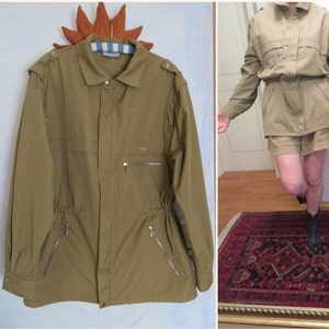 Könnte beinhalten: Eine khaki-grüne Utility-Jacke mit Kordelzug in der Taille und mehreren Reißverschlusstaschen. Die Jacke hat einen Kragen und lange Ärmel.