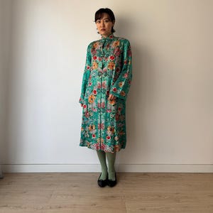 Op de afbeelding: Een lange, knoopsluitende jurk in een levendige teal kleur, versierd met een kleurrijk bloemenpatroon. De jurk heeft lange mouwen en een mandarijnkraag. Het model draagt groene panty's en zwarte ballerina's.