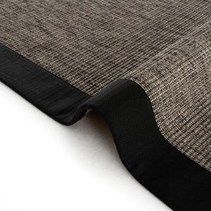 May include: Close-up of a woven rug with a textured, brown and beige surface. The rug has a thick, black border that curves at the corner. The rug is made of a durable material.