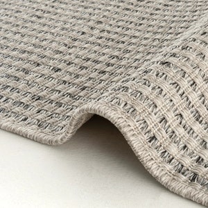 Op de afbeelding: Een geweven vloerkleed met een gestructureerd, rasterachtig patroon in tinten lichtgrijs, beige en donker antraciet. Het kleed heeft een dikke, afgewerkte rand en een dichte, verweven constructie die een uniform, geometrisch oppervlak vormt.