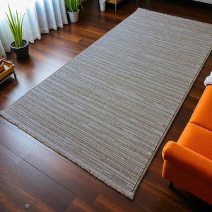 May include: A rectangular, neutral-toned area rug with a striped pattern. The rug is placed on a dark wood floor, and is near an orange sofa. The rug's dimensions are approximately 6 feet by 9 feet.
