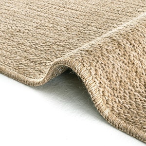 May include: A tan area rug with a dense, woven texture and a thick, stitched border. The rug is folded over at one corner, revealing the consistent, tight-knit construction of the natural-toned fibres throughout the entire surface area.