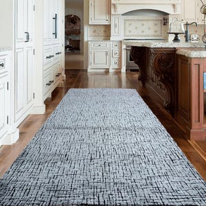 May include: A gray and white patterned area rug with a subtle geometric design. The rug is laid out in a kitchen with white cabinets and hardwood floors.
