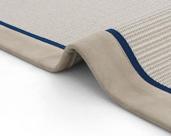 Puede incluir: Primer plano de una alfombra beige con ribete azul. La alfombra está doblada, mostrando el revés de la tela.