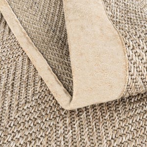 May include: Close-up of a woven rug in natural tones. The rug has a textured, patterned design with a folded edge in a lighter shade, creating a layered effect. The rug's fibres appear to be made of natural materials, suitable for a living space.