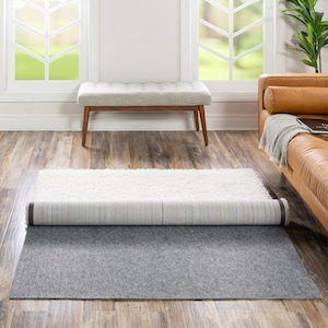 May include: A gray rug pad is rolled out on a wood floor. A white rug is rolled up on top of the rug pad. A wooden bench and a brown leather couch are in the background.
