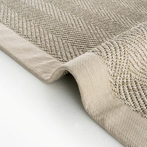 May include: A beige area rug featuring a woven herringbone pattern and a solid, smooth fabric border. The rug is folded at the corner, revealing the textured weave of the material and the consistent light tan color throughout the entire piece.