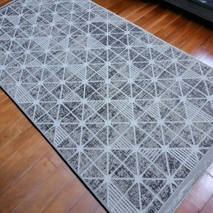 Puede incluir: Una alfombra de área gris y blanca con un patrón geométrico y un aspecto desgastado sutil. La alfombra tiene un patrón de diamante con líneas blancas sobre un fondo gris.