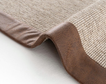Puede incluir: Primer plano de una alfombra beige tejida con un borde de cuero marrón. La textura de la alfombra es visible, y el borde de cuero está ligeramente curvado, mostrando sus detalles. La alfombra es de color neutro.