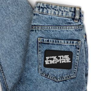 Op de afbeelding: Lichtblauwe spijkerbroek met een zwarte patch op de achterzak. De patch toont de tekst "F*ck The Empire" in witte blokletters. De jeans heeft een klassiek ontwerp met vijf zakken.