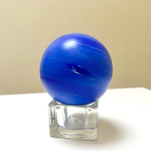 Puede incluir: Una esfera de vidrio azul con vetas blancas, que se asemeja a un planeta, se asienta sobre un cubo de vidrio transparente.