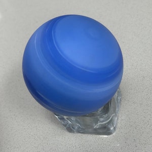 Puede incluir: Una esfera de vidrio azul con patrones blancos en espiral, sentada sobre un soporte de vidrio transparente.