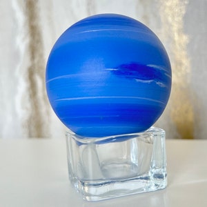 Puede incluir: Una esfera de vidrio azul con rayas blancas, que se asemeja a un planeta, se asienta sobre una base cuadrada de vidrio transparente.