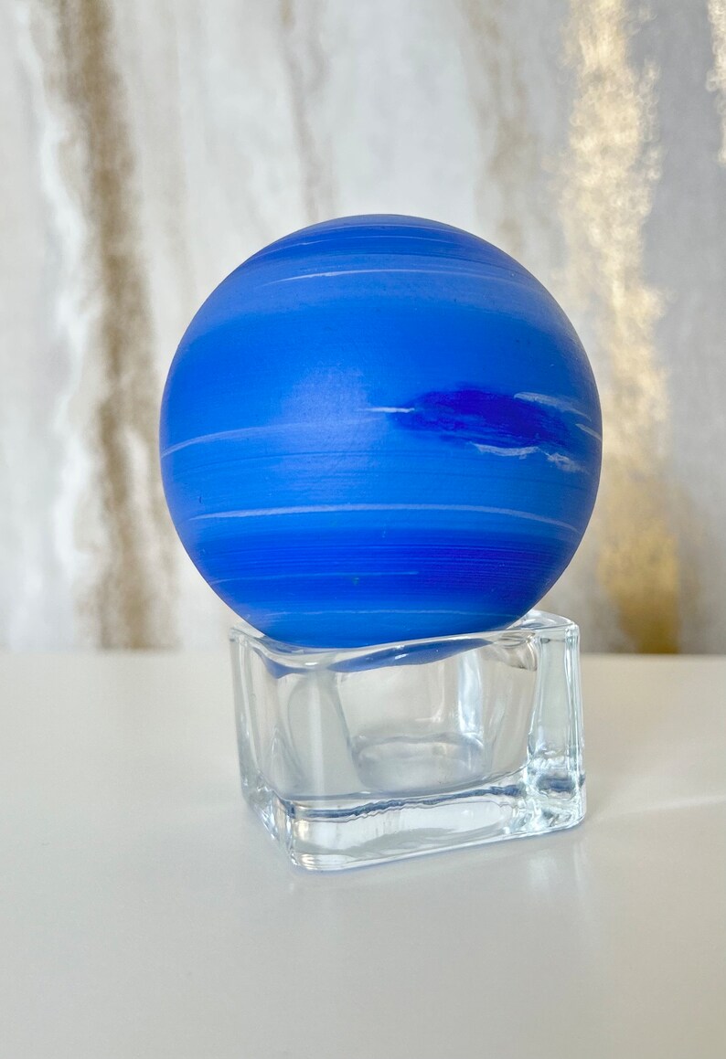 Puede incluir: Una esfera de vidrio azul con rayas blancas, que se asemeja a un planeta, se asienta sobre una base cuadrada de vidrio transparente.