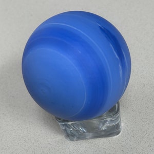Puede incluir: Una esfera de vidrio azul con remolinos blancos, que se asemeja a un planeta, sentada sobre un pedestal de vidrio transparente.