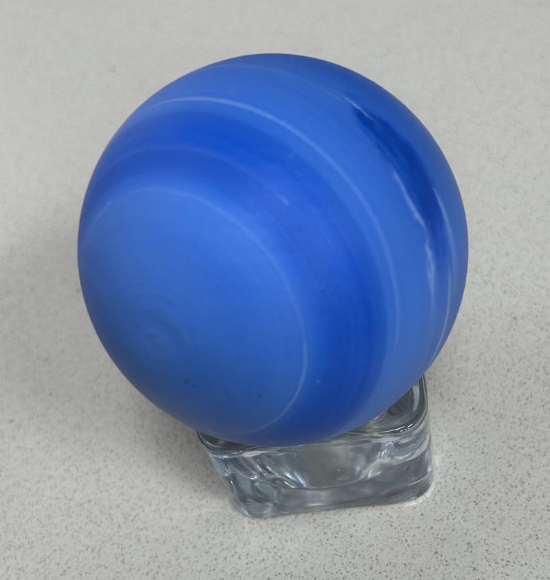 Puede incluir: Una esfera de vidrio azul con remolinos blancos, que se asemeja a un planeta, sentada sobre un pedestal de vidrio transparente.