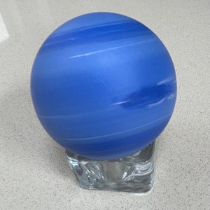 Puede incluir: Un globo de vidrio azul con rayas blancas, que se asemeja al planeta Neptuno, se encuentra sobre una base de vidrio transparente.