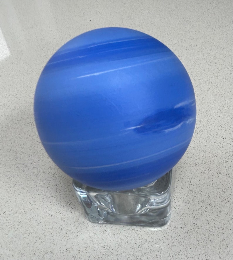 Puede incluir: Un globo de vidrio azul con rayas blancas, que se asemeja al planeta Neptuno, se encuentra sobre una base de vidrio transparente.