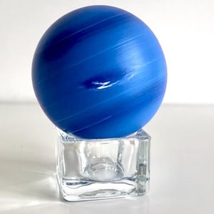 Puede incluir: Una esfera de vidrio azul con vetas blancas, que se asemeja a un planeta, se asienta sobre un soporte de vidrio transparente en forma de cubo.