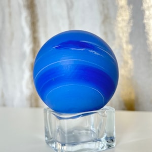 Puede incluir: Una esfera de vidrio azul con rayas blancas, que se asemeja a un planeta, se asienta sobre un cubo de vidrio transparente.