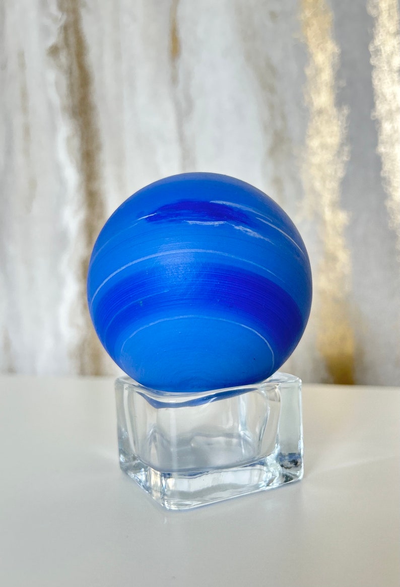 Puede incluir: Una esfera de vidrio azul con rayas blancas, que se asemeja a un planeta, se asienta sobre un cubo de vidrio transparente.