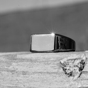 May include: A silver signet ring with a rectangular top. The ring is sitting on a wooden surface.