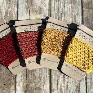 May include: Four crocheted cup cozies in red, orange, yellow, and mustard yellow. Each cozy is made with love and has a tag that says "Cup Cozy" and "Made with love".