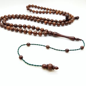 Peut inclure: Un chapelet en bois avec des perles marron foncé et un pendentif allongé assorti. Les perles sont enfilées sur un cordon vert. Le chapelet est présenté sur un fond blanc.