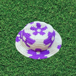 Op de afbeelding: Een gehaakte bucket hat met een witte basis en paarse bloemvormige ontwerpen.