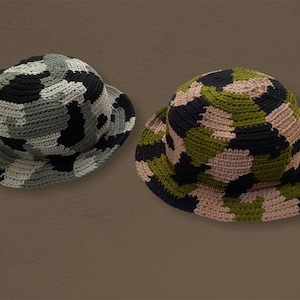 以下が含まれることがあります： 迷彩柄のクロシェの バケットハットが2つ。1つは緑、黒、ベージュ、もう1つは緑、黒、茶色です。