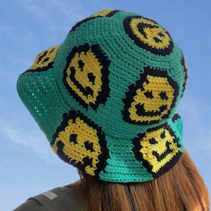 May include: A green crocheted bucket hat with yellow smiley faces. The hat is made of a soft, textured yarn and has a wide brim.
