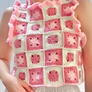 May include: A pink and white crochet top with a ruffled neckline. The top is made of granny squares with a floral pattern.