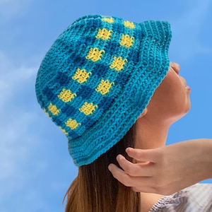 Może przedstawiać: Niebieski, szydełkowy kapelusz typu bucket z żółtymi kropkami. Kapelusz nosi osoba z długimi brązowymi włosami.