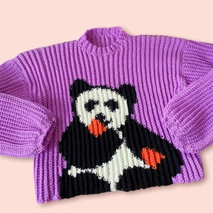 May include: A purple crocheted sweater with a black and white panda design. The panda is holding a red and orange fruit.