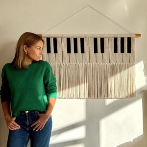 Peut inclure: Une suspension murale en macramé blanc et noir avec un motif de touches de piano. La suspension a une bordure en frange et est montée sur une baguette en bois.
