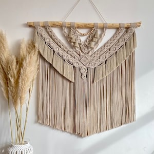 May include: A beige macrame wall hanging with a geometric design and wooden dowel. The hanging has a fringe border and is decorated with wooden beads.