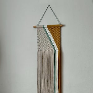 Puede incluir: Un tapiz de macramé con un diseño geométrico. La sección superior presenta un patrón tejido de color crema con una franja diagonal amarilla mostaza y verde. La parte inferior tiene una larga franja beige. Cuelga de una clavija de madera.