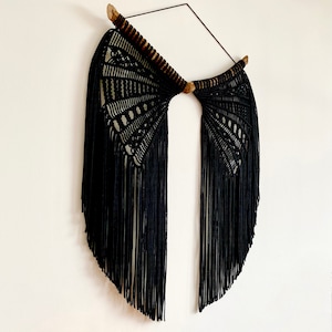 Puede incluir: Colgante de pared de macramé negro con listones de madera y flecos, colgado en una pared blanca con tres calabazas naranjas en primer plano.