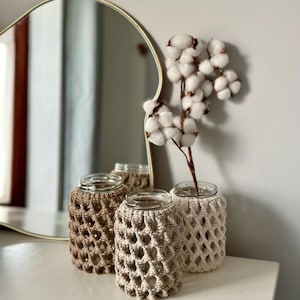 Peut inclure: Trois bocaux en verre avec des couvercles en crochet beige et marron. Les bocaux sont disposés sur une surface blanche avec une branche de coton en arrière-plan.