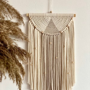 May include: A beige and white macrame wall hanging with a half-circle design and wooden beads. The hanging is made with thick cotton cord and has a natural, bohemian style.