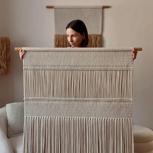 May include: A beige macrame wall hanging with a wooden dowel. The handmade textile features intricate knotting and vertical fringe. A smaller, similar wall hanging is visible in the background, along with a woman holding the larger piece.