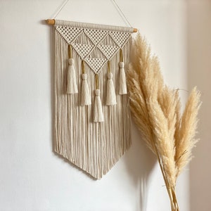 Può includere: Un arazzo da parete in macramè beige con un motivo geometrico e frange. L'arazzo è montato su una parete bianca con un mazzo di erba della pampa secca a destra.