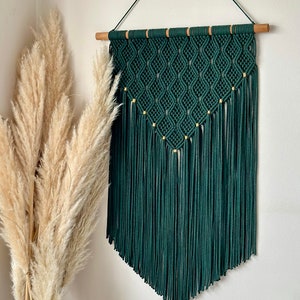 Puede incluir: Un tapiz de macramé de color verde oscuro con cuentas doradas y una barra de madera. El tapiz tiene un patrón de diamante y flecos.